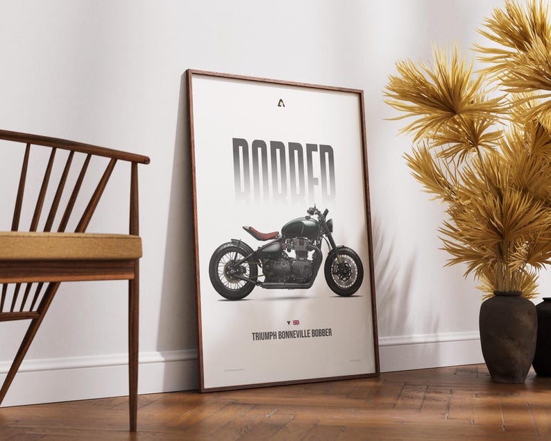 May include: Framed art print featuring a dark green Triumph Bonneville Bobber motorcycle. The word "BOBBER" is printed in large, bold, gray letters above the motorcycle. The print is displayed on a white background with a wooden frame.