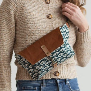May include: A crossbody bag featuring a dark teal fabric with a white repeating wave pattern and a solid brown flap. The bag includes a light brown leather strap secured with a metal snap and a brass-toned swivel clasp for closure and attachment.