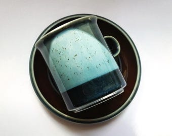 May include: A ceramic mug and saucer set featuring a two-tone glaze. The mug has a speckled light blue upper section and a solid dark blue base, while the saucer is a deep brown with a dark blue rim, viewed from a top-down perspective.