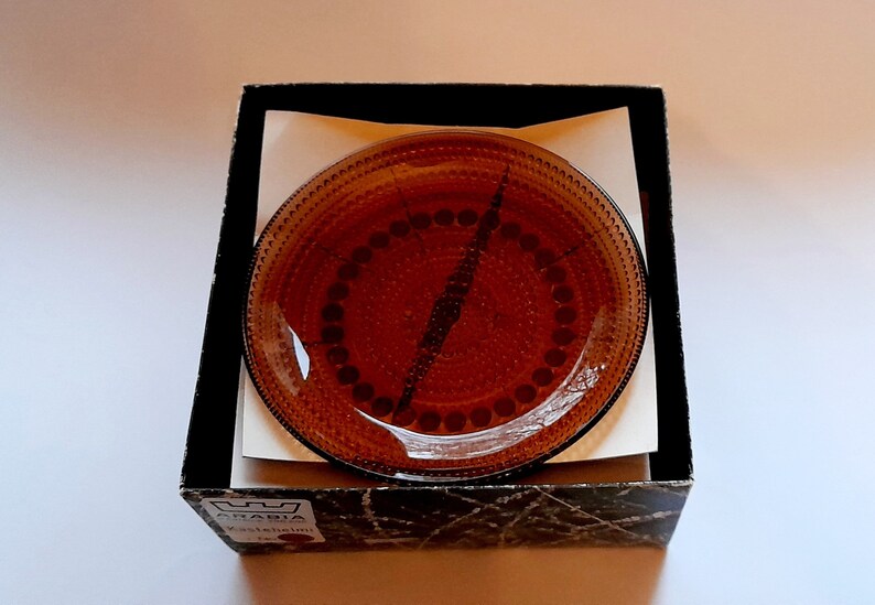 May include: A brown glass plate with a decorative pattern of small circles and dots. The plate is in a black box with the word "ARABIA" printed on the bottom.
