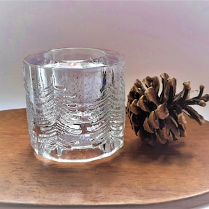 May include: A clear glass candle holder with a textured design of pine trees. The candle holder is sitting on a wooden surface next to a brown pine cone.