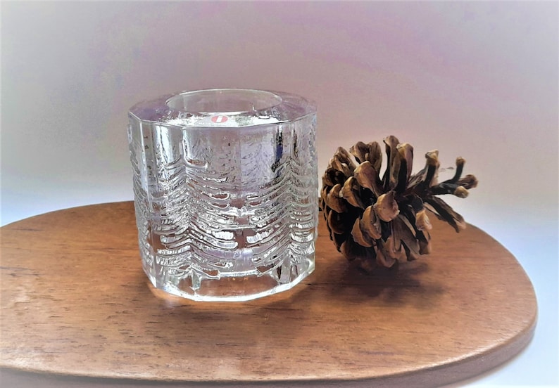 May include: A clear glass candle holder with a textured design of pine trees. The candle holder is sitting on a wooden surface next to a brown pine cone.