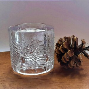 May include: A clear glass candle holder with a textured design resembling a forest of trees. The holder is sitting on a wooden surface next to a brown pine cone.