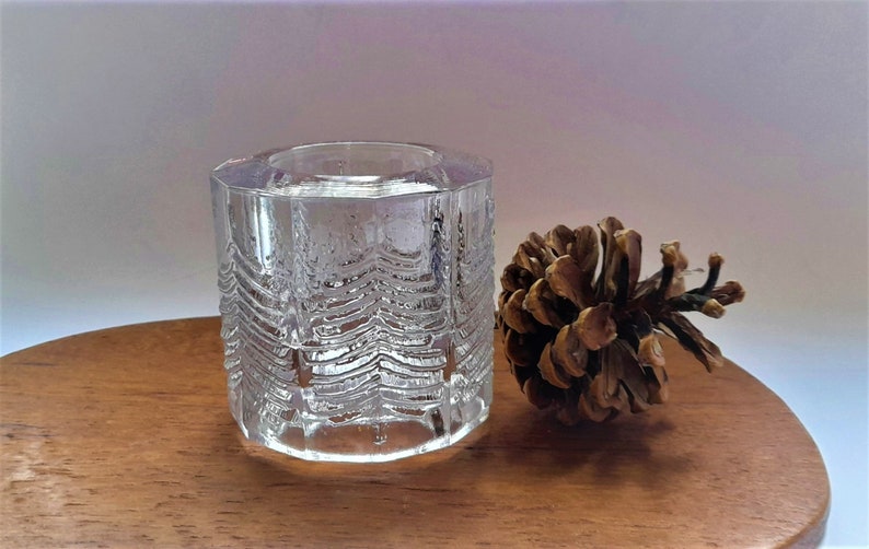 May include: A clear glass candle holder with a textured design resembling a forest of trees. The holder is sitting on a wooden surface next to a brown pine cone.