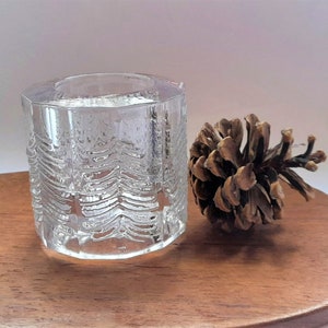 May include: A clear glass candle holder with a textured design resembling pine trees. The holder is sitting on a wooden surface next to a brown pine cone.