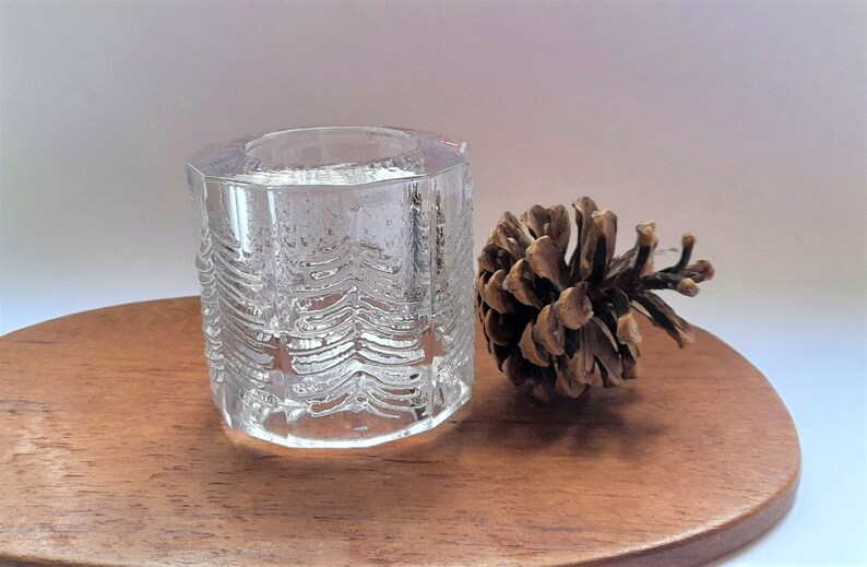 May include: A clear glass candle holder with a textured design resembling pine trees. The holder is sitting on a wooden surface next to a brown pine cone.