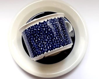 May include: A ceramic cup and saucer set featuring a white base color with a dense, repeating pattern of small dark blue flowers covering the exterior of the cup. The saucer has a solid dark blue center surrounded by a wide, plain white rim.