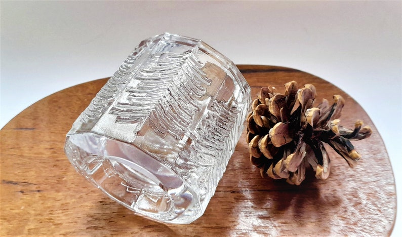 May include: A clear glass candle holder with a textured design of pine trees. The holder is sitting on a wooden surface next to a pine cone.