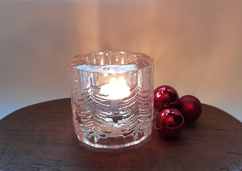 May include: A clear glass candle holder with a textured design of Christmas trees. The holder is lit with a candle and surrounded by three red ornaments.