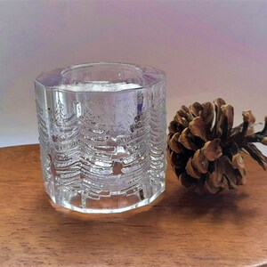 May include: A clear glass candle holder with a textured design of trees. The holder is sitting on a wooden surface next to a brown pine cone.