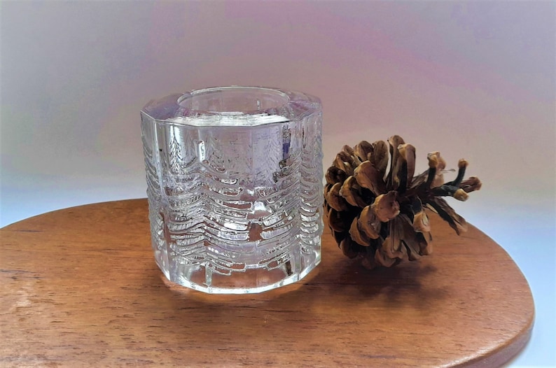 May include: A clear glass candle holder with a textured design of trees. The holder is sitting on a wooden surface next to a brown pine cone.