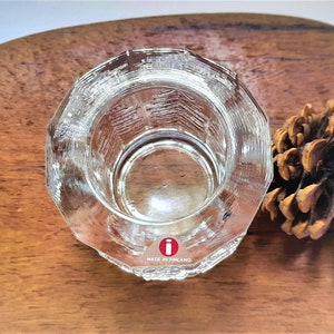 May include: A clear glass candle holder with a textured design. The holder has a round base and a cylindrical shape. The bottom of the holder has a red "i" logo and the text "Made in Finland".