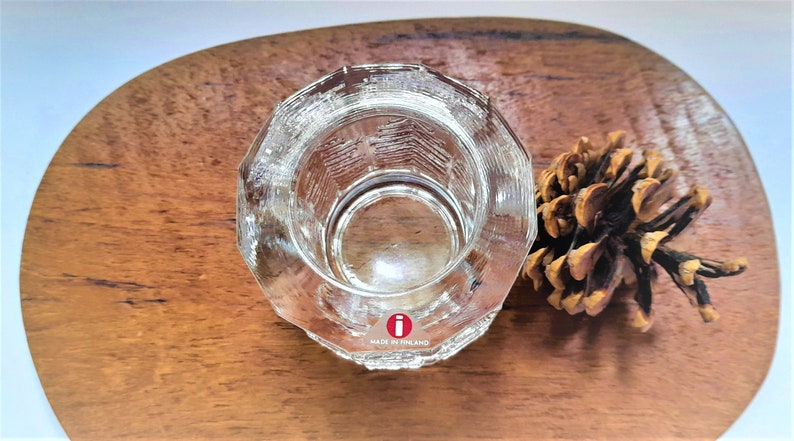 May include: A clear glass candle holder with a textured design. The holder has a round base and a cylindrical shape. The bottom of the holder has a red "i" logo and the text "Made in Finland".