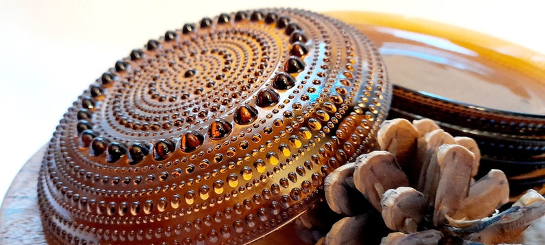 May include: A set of amber glass plates with a textured, dotted surface. The plates are stacked on top of each other and a pine cone is in the foreground.