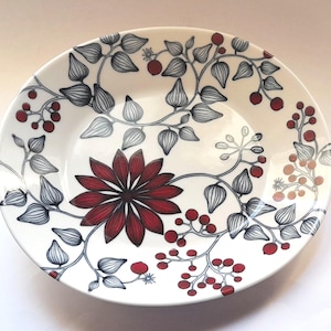May include: A white ceramic bowl featuring a central dark red flower surrounded by intricate black line-drawn vines, leaves, and clusters of small red berries. The botanical pattern extends across the interior surface of the round dish.