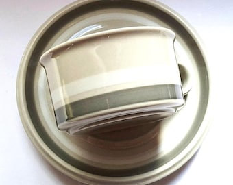 May include: A ceramic cup and saucer set featuring a smooth, glossy finish in neutral tones. The design includes horizontal bands of light beige, cream, and a darker muted gray-green color that wraps around both the cup and the circular saucer.