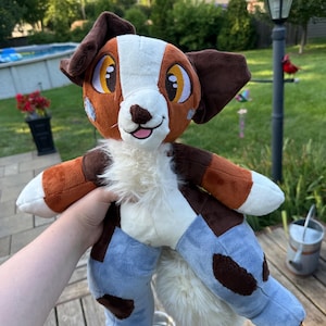 May include: A plush dog toy with brown, white, and blue accents. The toy has large, expressive eyes, floppy ears, and a fluffy tail. The dog is wearing blue pants with brown spots.