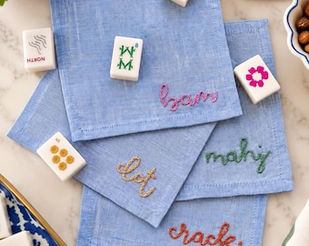 May include: Four square light blue linen napkins, each featuring a unique word embroidered in a different colored thread: pink, yellow, green, and orange. The napkins are arranged with scattered white mahjong tiles featuring various symbols.
