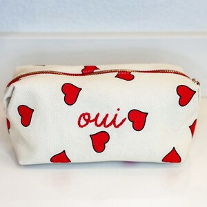 May include: White canvas cosmetic bag with a red zipper and red heart pattern. The word "oui" is embroidered in red on the front. The bag is rectangular and appears to be made of a sturdy material.