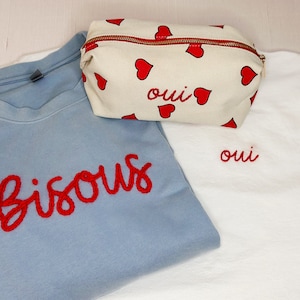 May include: A light blue sweatshirt with the word "Bisous" embroidered in red, a white t-shirt with the word "oui" embroidered in red, and a cream-colored cosmetic bag with red hearts and the word "oui".