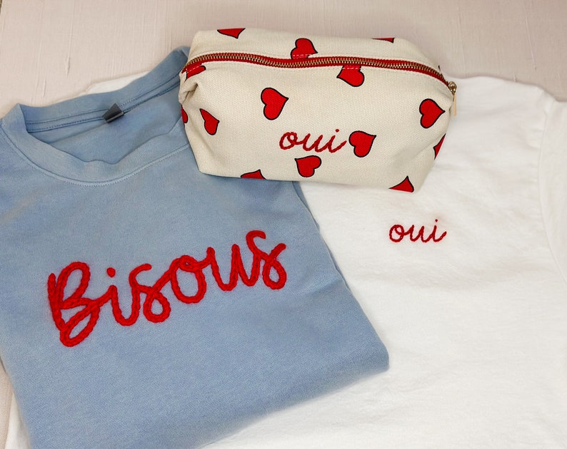 May include: A light blue sweatshirt with the word "Bisous" embroidered in red, a white t-shirt with the word "oui" embroidered in red, and a cream-colored cosmetic bag with red hearts and the word "oui".