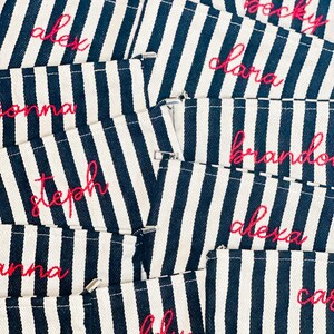 May include: Close-up of several navy and white striped pouches with names embroidered in pink thread. Names include Angie, Alex, Becky, Clara, Brandon, Alexa, Steph, Anna, Addy, and Cara.