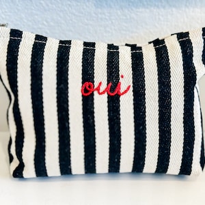 May include: A small, rectangular black and white striped pouch with a zipper closure. The word "oui" is embroidered in red on the front. The pouch is made of a woven fabric.