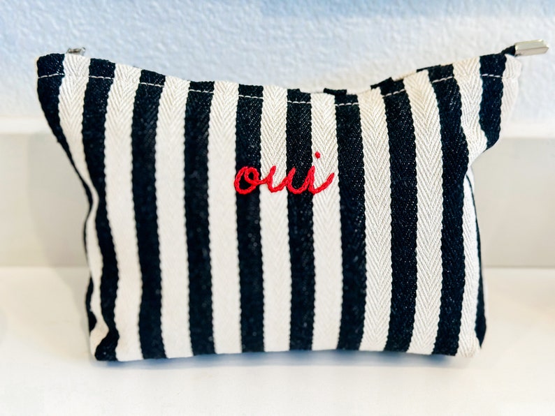 May include: A small, rectangular black and white striped pouch with a zipper closure. The word "oui" is embroidered in red on the front. The pouch is made of a woven fabric.