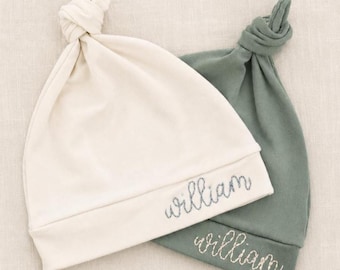 May include: Two baby knot hats are shown, one cream and one sage green. The cream hat has the name "william" embroidered in blue thread. The sage green hat also has the name "william" embroidered in blue thread. Both hats are made of soft fabric.
