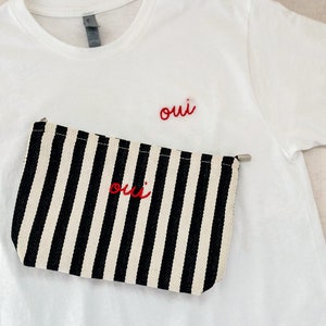 May include: A white t-shirt with the word "oui" embroidered in red. A black and white striped pouch with the word "oui" in red is placed on the shirt.