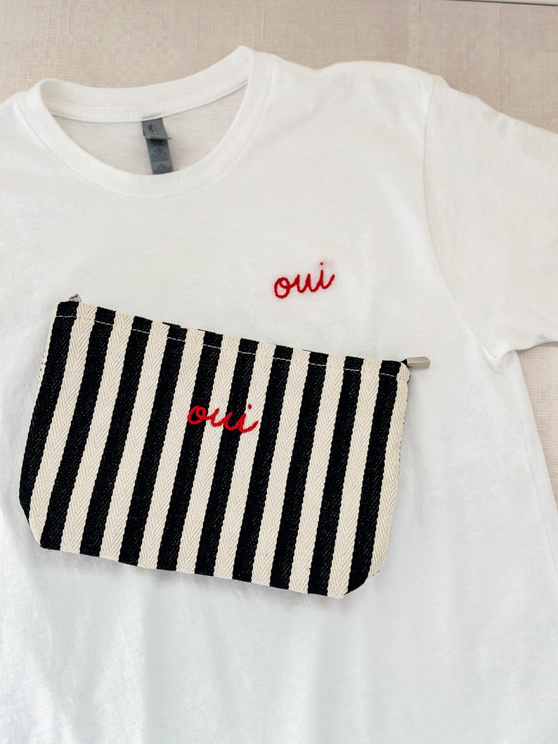 May include: A white t-shirt with the word "oui" embroidered in red. A black and white striped pouch with the word "oui" in red is placed on the shirt.