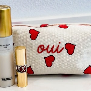 May include: A cosmetic set featuring a white and red heart-patterned makeup bag with the word "oui" in red, a gold-capped spray bottle labeled "Boum-Boum Milk", and a gold lipstick tube.