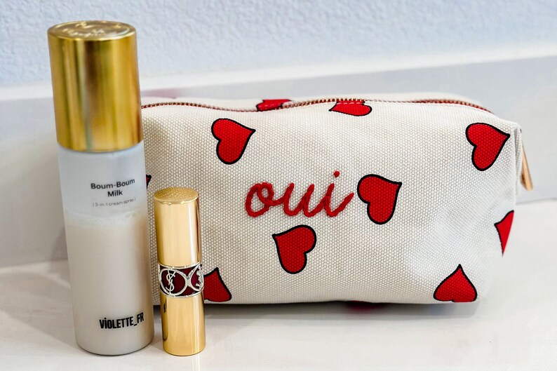 May include: A cosmetic set featuring a white and red heart-patterned makeup bag with the word "oui" in red, a gold-capped spray bottle labeled "Boum-Boum Milk", and a gold lipstick tube.