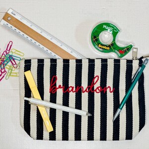 May include: A black and white striped pencil case with the name "Brandon" embroidered in red. A wooden ruler, tape dispenser, pens, and paper clips are also visible.