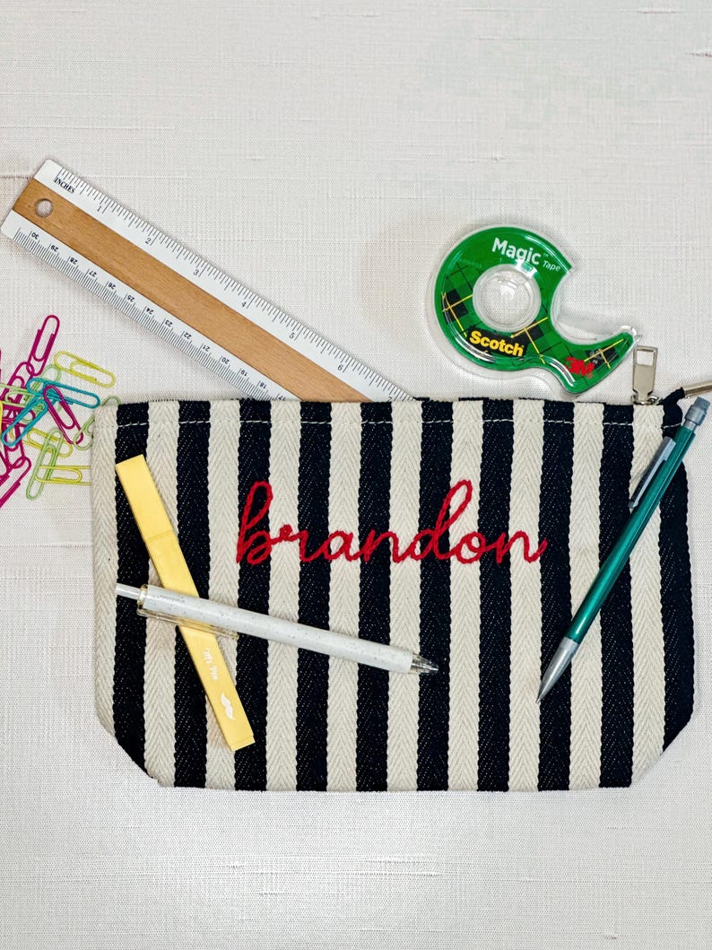 May include: A black and white striped pencil case with the name "Brandon" embroidered in red. A wooden ruler, tape dispenser, pens, and paper clips are also visible.