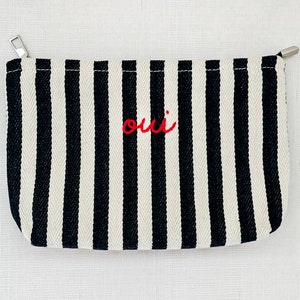 May include: A black and white striped pouch with a silver zipper. The word "oui" is embroidered in red on the front. The pouch is rectangular and appears to be made of canvas or a similar fabric.