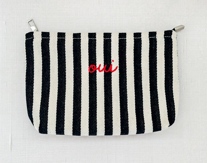 May include: A black and white striped pouch with a silver zipper. The word "oui" is embroidered in red on the front. The pouch is rectangular and appears to be made of canvas or a similar fabric.