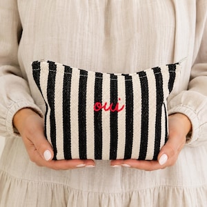 May include: A black and white striped cosmetic bag with the word "oui" embroidered in red. The bag has a zipper closure and is held by a person wearing a beige dress.