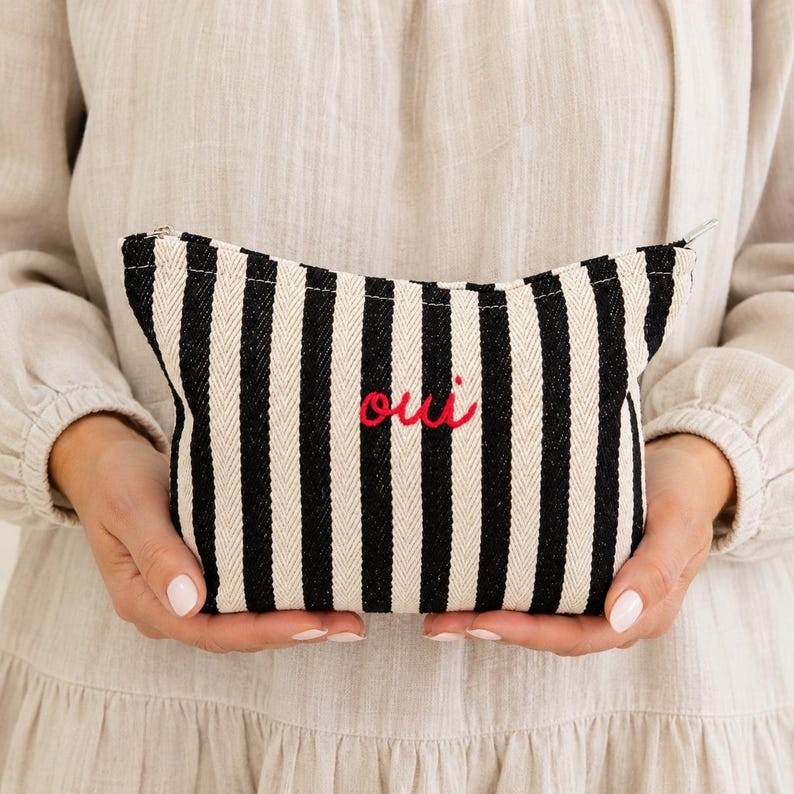 May include: A black and white striped cosmetic bag with the word "oui" embroidered in red. The bag has a zipper closure and is held by a person wearing a beige dress.