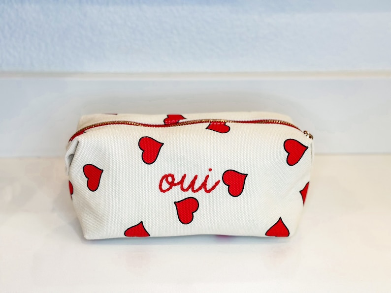 May include: White canvas cosmetic bag with a red zipper and red heart pattern. The word "oui" is embroidered in red on the front. The bag is rectangular and appears to be made of a sturdy material.