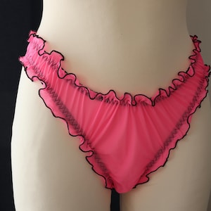 May include: Bright pink underwear with a ruffled edge and black trim. The sheer fabric is gathered at the waist and leg openings. The garment is displayed on a mannequin.