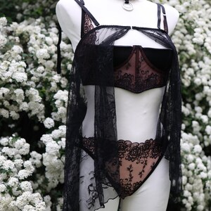 Puede incluir: Conjunto de lencer&iacute;a negra sobre un maniqu&iacute; blanco, con sujetador, bragas y una cubierta transparente. El sujetador y las bragas tienen detalles de encaje floral. Un collar&iacute;n negro con un anillo plateado est&aacute; alrededor del cuello del maniqu&iacute;.