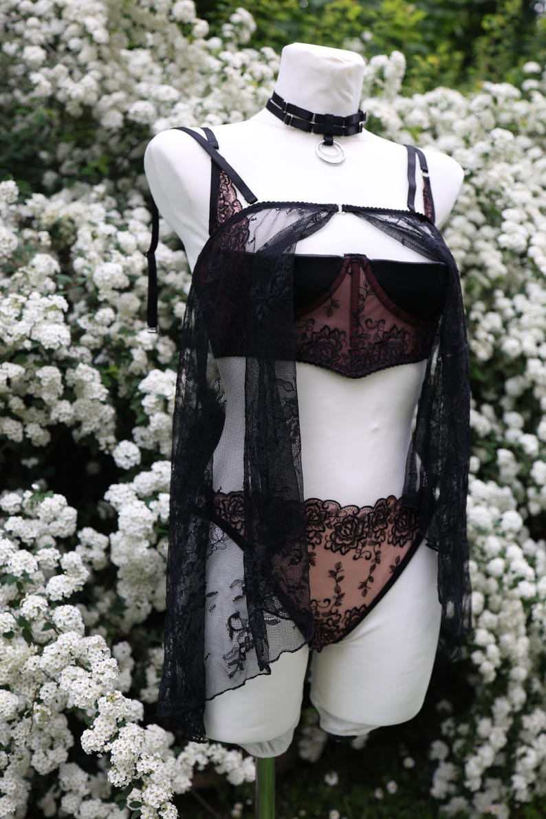 Puede incluir: Conjunto de lencer&iacute;a negra sobre un maniqu&iacute; blanco, con sujetador, bragas y una cubierta transparente. El sujetador y las bragas tienen detalles de encaje floral. Un collar&iacute;n negro con un anillo plateado est&aacute; alrededor del cuello del maniqu&iacute;.