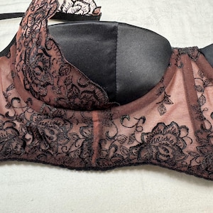 Puede incluir: Sujetador negro y color rubor con intrincados detalles de encaje floral. El sujetador presenta un acabado satinado en las copas y delicados detalles de encaje a lo largo de los bordes, creando un aspecto rom&aacute;ntico y elegante.