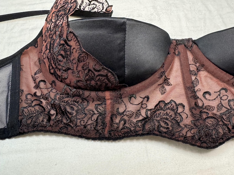 Puede incluir: Sujetador negro y color rubor con intrincados detalles de encaje floral. El sujetador presenta un acabado satinado en las copas y delicados detalles de encaje a lo largo de los bordes, creando un aspecto rom&aacute;ntico y elegante.