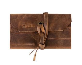 May include: A brown leather wallet with a knotted strap and a metal button. The wallet has the text "Tanner Goods" embossed on the front.