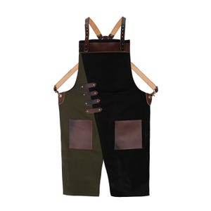 Puede incluir: Delantal bicolor en verde oliva oscuro y negro, con detalles de cuero marrón en bolsillos, correas y hebillas. El delantal tiene correas cruzadas. El diseño es dividido, creando una estética única. Adecuado para uso culinario o artesanal.