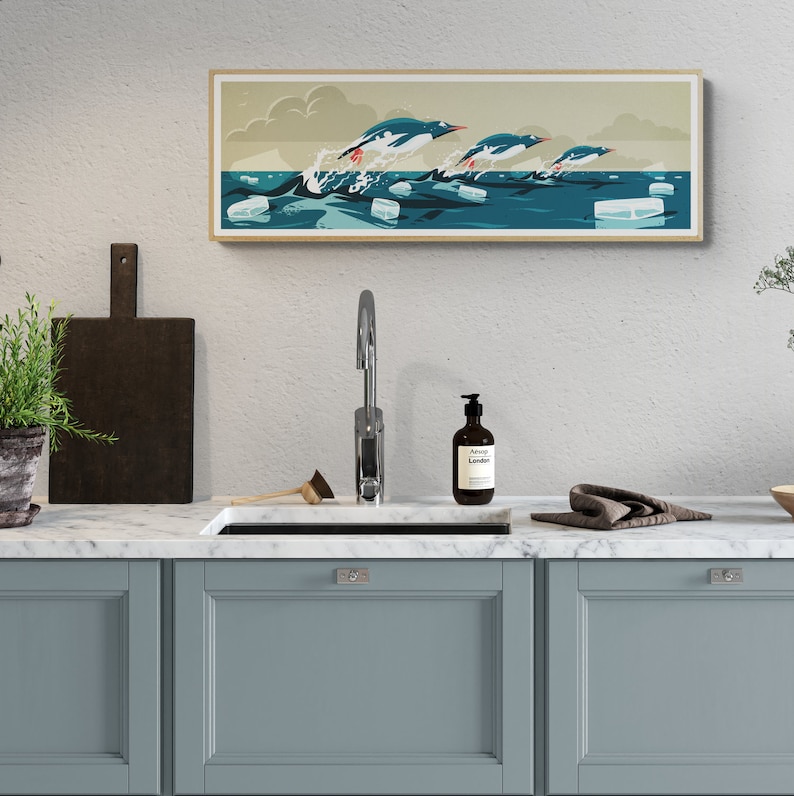 Long horizontal poster or art print hanging in a kitchen with artwork of three gentoo penguins that are making speed between the ice floes in the ocean around Antarctica. The panorama has shades of beige, white and blue colors.