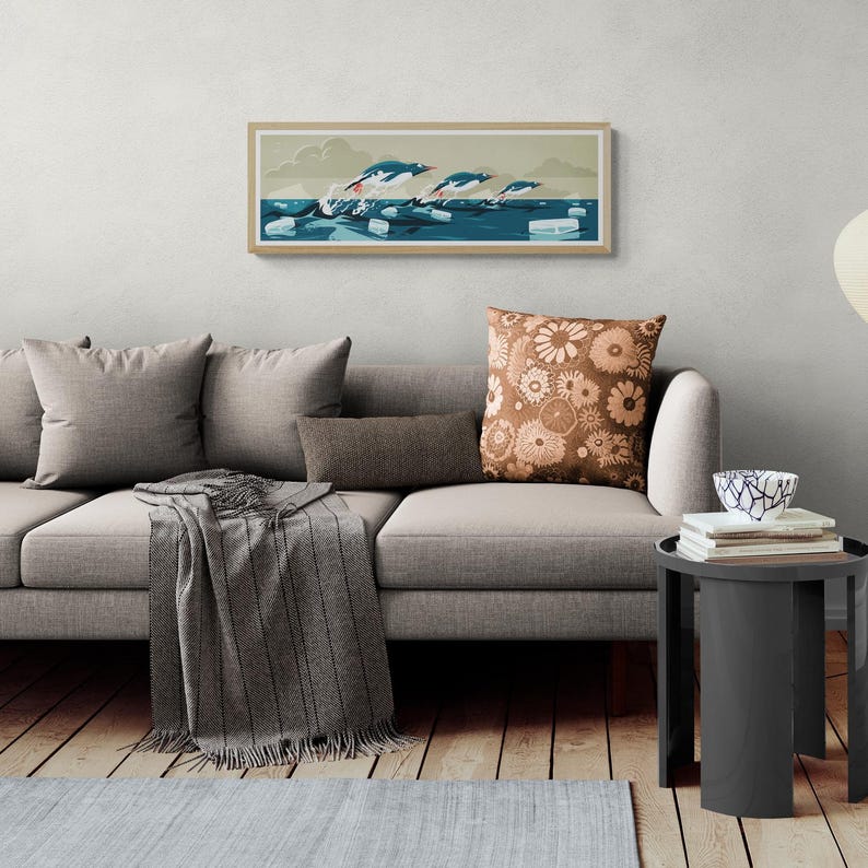 Long narrow horizontal poster or art print hanging in a living room with artwork of three gentoo penguins that are making speed between the ice floes in the ocean around Antarctica. The panorama has shades of beige, white and blue colors.
