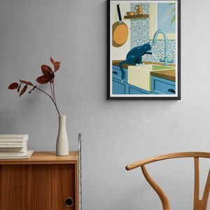 Living room with a poster on the wall. The illustration on the art print shows a black cat in a kitchen with azulejo tiles as can often be found in Spain and Portugal. The artwork gives a Mediterranean feel. The cat is playing with dripping water.